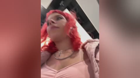 Media: Video of a young woman with vibrant red hair, wearing a pink top and a silver necklace, standing in a dimly lit room with blurred figures in the background.