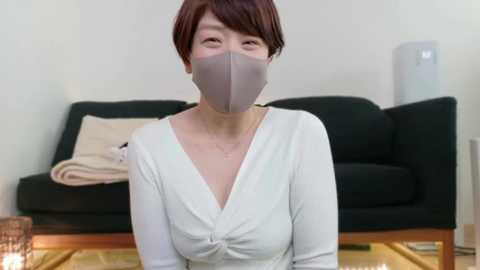 Media: Video of an Asian woman with short brown hair, wearing a grey face mask and a white V-neck top, seated on a wooden floor in a minimalist living room with a black sofa and a cream throw pillow.