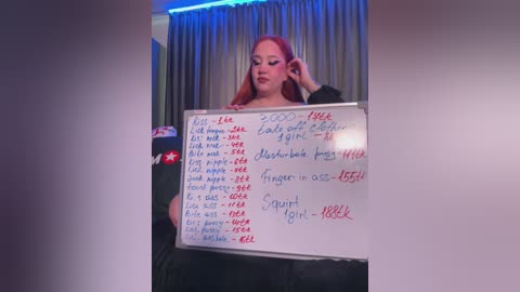 Media: Video of a woman with long red hair, holding a whiteboard listing BDSM terms, set in a dimly lit room with blue curtains.