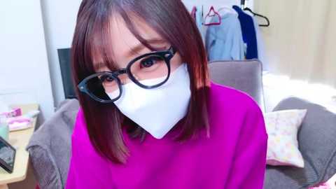 Media: Video of a young East Asian woman with straight, shoulder-length brown hair, wearing black-framed glasses, a pink sweater, and a white mask, sitting in a cozy living room with a TV, clothes hanging, and a gray couch.