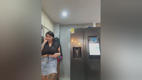 Media: A video of a Latina woman with long black hair, medium build, wearing a black crop top and denim skirt, talking on a cell phone in a modern, dimly lit hallway with gray walls and a fridge.