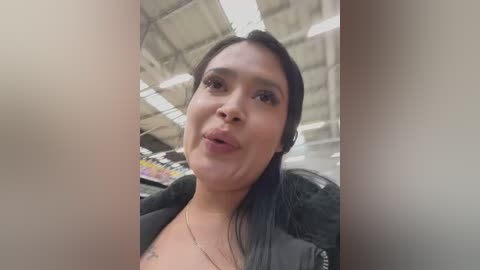 Media: Video of a young Latina woman with medium skin tone and long black hair, wearing a black jacket and a necklace. She stands in a supermarket aisle with fluorescent lighting and visible shelves in the background.