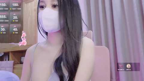 Media: Video of an Asian woman with long black hair, wearing a white face mask, light gray spaghetti strap top, sitting indoors with a wooden door and pink flowers in the background.