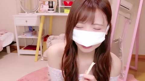 Media: Video of a young Asian woman with long brown hair, wearing a white mask and a light pink off-shoulder top, sitting on a pink rug in a cozy, softly lit bedroom with white furniture.