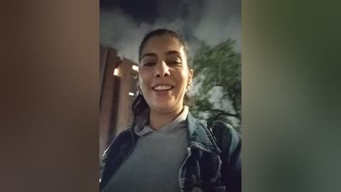 Media: Video of a young woman with dark hair tied back, smiling broadly, wearing a grey shirt and denim jacket, standing outdoors in a dimly lit, foggy evening with greenery and a building in the background.