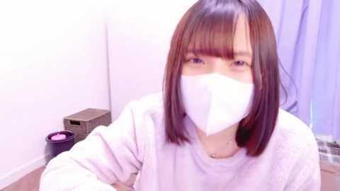 Media: Video of an East Asian woman with straight brown hair and bangs, wearing a white face mask and a white sweater, in a simple room with a wicker basket and a pink candle.