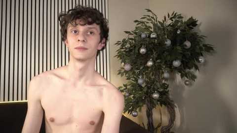 Video of a thin, shirtless young man with curly hair, standing in front of a fake Christmas tree with silver ornaments and a beige wall with vertical black stripes.