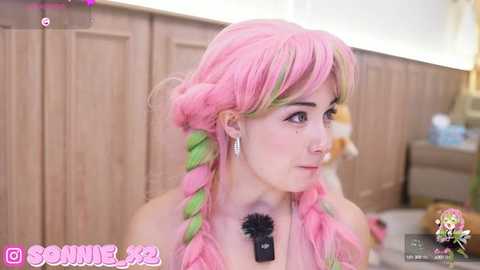 Media: Video of a young woman with pink hair in braids, wearing a black top with a microphone, standing in a kitchen with wooden cabinets.