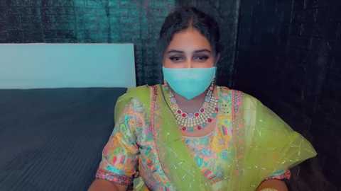 Media: Video of a young South Asian woman wearing a colorful, floral-patterned sari, a blue mask, and heavy jewelry, seated indoors with a dark wall and white baseboard in the background.