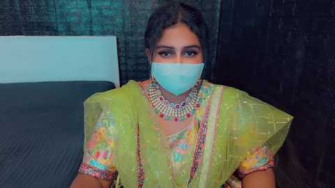 Media: Video of a South Asian woman with dark skin, wearing a light green sari with intricate embroidery, a pink and gold necklace, and a light blue face mask, sitting on a black cushioned surface.