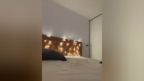 Media: Video of a minimalist bedroom with a beige bed, a wooden headboard adorned with warm, glowing string lights, and a closed white door in the background.