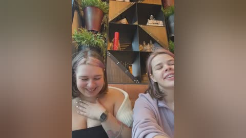 Media: Video of two young women laughing, one in a black off-shoulder top, the other in a light sweater, against a backdrop of a modern, wooden wall with green potted plants and decorative items.