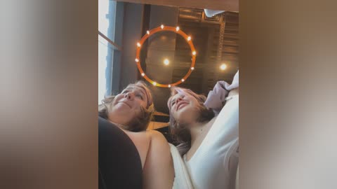 Media: Video of two women with blonde hair, one in a black top, the other in white, lying on a couch, gazing upwards at a circular, illuminated light fixture, creating a cozy, intimate indoor setting.