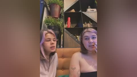 Media: Video of two young women, one with pink hair, the other with dark hair, sitting on a brown couch in a dimly lit room with green plants and a black shelving unit.