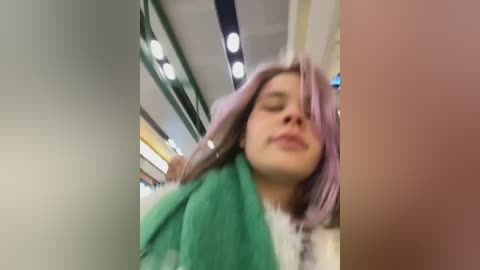 Media: Video of a person with lavender hair, wearing a white jacket, leaning against a green towel in a brightly lit indoor setting with overhead lights and blurred background.