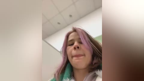 Media: Video of a young girl with light skin, straight pink hair, and a slight smile. She wears a green and white patterned shirt in a classroom with white ceiling tiles and fluorescent lights.