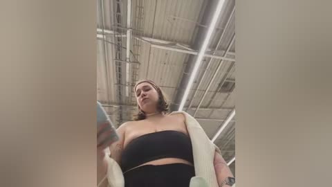 Media: Video of a plus-sized woman with medium skin tone and shoulder-length brown hair, wearing a black bandeau top and black leggings. She holds a smartphone, standing in an industrial warehouse with exposed ceiling beams and fluorescent lighting.