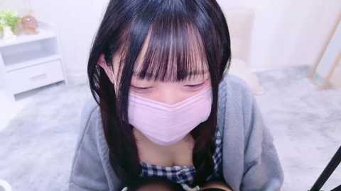 Media: Video of an Asian woman with long black hair and pale skin, wearing a pink face mask, a gray cardigan, and a checkered top, sitting on a light-colored floor in a minimalistic room.