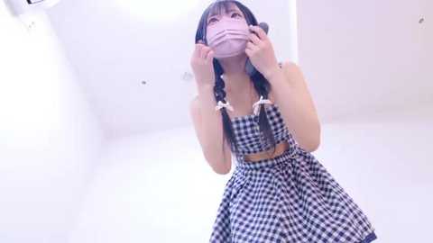 Media: Video of an Asian woman in a black-and-white checkered dress, wearing a pink face mask, holding it up, indoors with a white background.