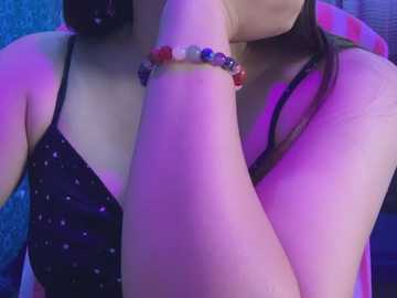 Media: A close-up video of a young woman with light skin, wearing a black spaghetti-strap top with small white polka dots. She has a colorful beaded bracelet on her right wrist, and her long dark hair partially covers her face. The background is a mix of blue and purple lighting.