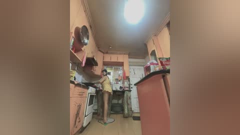 Media: A video of a small, cluttered kitchen with a person in yellow shorts and a white shirt standing near the stove, surrounded by cabinets and countertops.