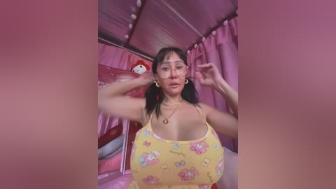 Media: Video of an East Asian woman with large breasts, wearing a yellow floral camisole, adjusting her hair, in a pink-themed room with plush toys.