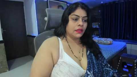 Media: A video shows a middle-aged South Asian woman with medium skin tone, dark hair, and red lipstick, wearing a white lace top and blue sari, seated in a modern, dimly lit room with a TV and a bed in the background.