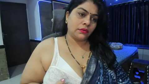 Media: Video of a South Asian woman with medium skin tone, long black hair, wearing a white lace bra and a blue patterned saree. She sits in a modern room with dark curtains and a bed in the background.