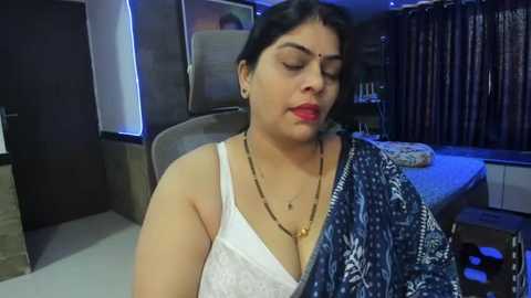 Media: A video of an Indian woman with medium complexion, wearing a white lace bra and a blue patterned sari, sitting in a modern living room with wooden furniture, bookshelves, and a bed in the background.