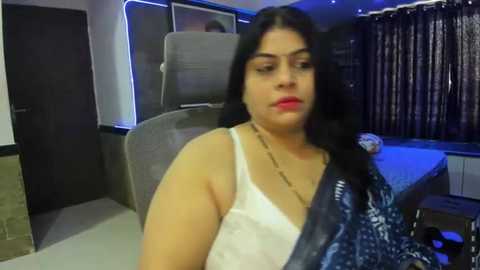 Media: A video of a South Asian woman with long black hair, fair skin, and wearing a white lace bra and blue patterned sari, sitting in a modern office with grey chairs and dark curtains.