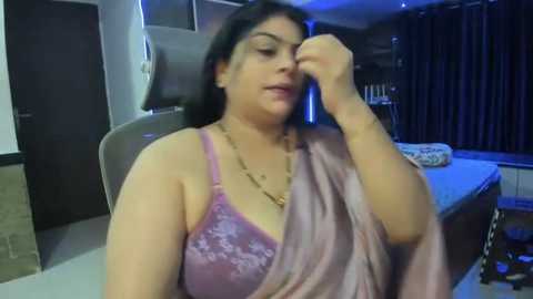 Media: Video of a South Asian woman with dark hair, wearing a pink satin blouse, adjusting her hair in a dimly lit room with dark curtains, a door, and a table.