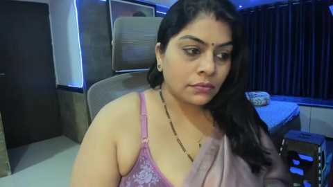 Media: Video of a South Asian woman with medium skin tone, long dark hair, wearing a pink lace bra and beige sari, sitting in a dimly-lit room with modern furniture and dark curtains.