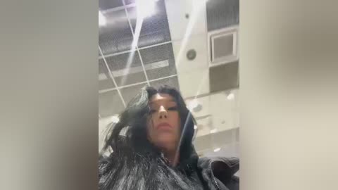 Media: A video of a young woman with medium-brown skin, long black hair, and a grey hoodie, captured from a low angle inside a modern room with a tiled ceiling and fluorescent lights.