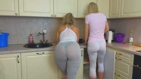 Media: Video of two women in tight white leggings and light pink tops, leaning over a kitchen sink, showcasing their curvy, ample buttocks.