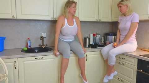 Media: Video of two middle-aged women, one blonde, the other with brown hair, sitting on kitchen counters in matching light pink athletic attire, discussing.