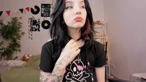 Media: Video of a pale-skinned, black-haired woman with tattoos and piercings, wearing a black T-shirt with a cartoon skull design. She stands in a cozy bedroom with a green bed, bamboo plant, and vinyl records on the wall.