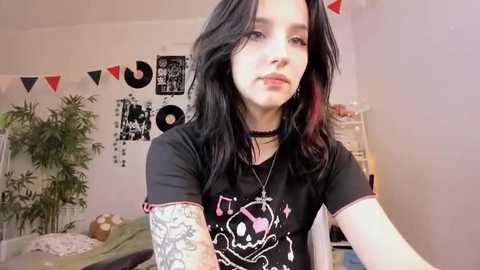 Media: A young woman with long black hair, pale skin, and tattoos poses in a cozy bedroom with a black tee, choker, and a vinyl record-themed banner.