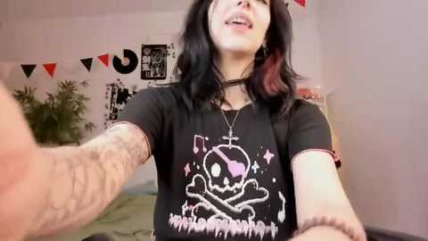 Media: A video shows a pale-skinned woman with long black hair, wearing a black t-shirt with a skull graphic, smiling in a room with black and white decor.