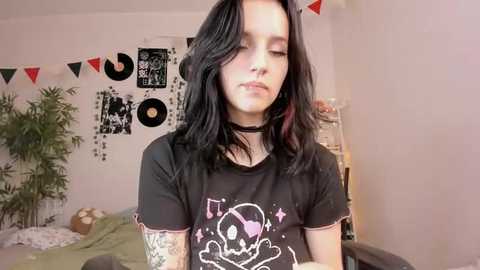 Media: Video of a pale-skinned young woman with black hair, wearing a black graphic t-shirt, black choker, and tattoos, standing in a bedroom with a wall adorned with records, posters, and bunting.