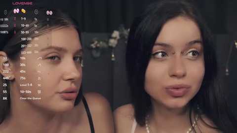 Media: Video of two young women with fair skin, dark hair, and light makeup, smiling at the camera. Background shows a dimly lit room with a TV displaying a live stream.