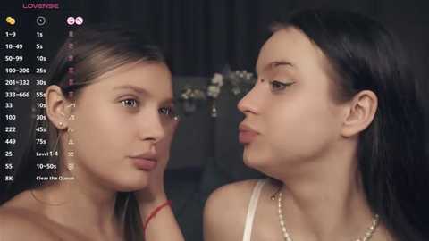 Media: Video of two young women with light skin and long dark hair, wearing minimal makeup, captured in a close-up indoor setting with a blurred background.