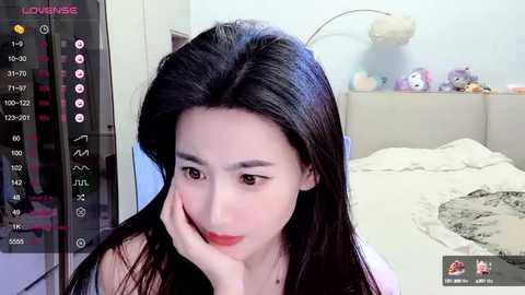 Media: Video of a young Asian woman with long black hair, wearing a sleeveless top, resting her head on her hand, in a bedroom with a bed, stuffed toys, and a camera screen.