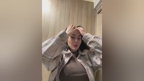 Media: A video shows a woman with fair skin, straight brown hair, and a plaid shirt, running her hands through her hair. She stands in a beige-walled room with wooden paneling.