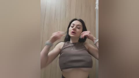 Media: A video of a young woman with fair skin and dark hair, wearing a brown crop top, adjusting her hair. She has a slender physique and small breasts. The background is a beige wooden wall.