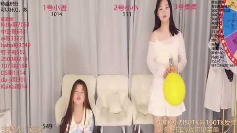 Media: Video of two Asian women in white dresses, one holding a yellow balloon, standing in front of a white couch and gray curtains. Text overlays in Chinese characters.