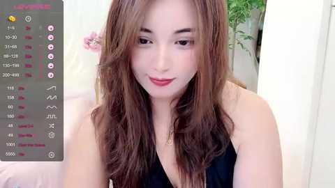 Media: A video of an Asian woman with long, wavy brown hair, fair skin, and red lipstick, wearing a black sleeveless top. Background shows a white wall and green plant.