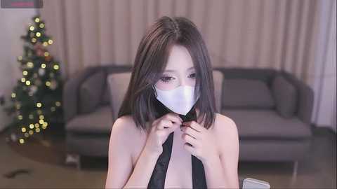 Media: A video of an Asian woman with shoulder-length black hair, wearing a white mask, adjusting her black strapless top in a modern, beige-furnished room with a Christmas tree and sofa.