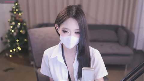 Media: A video of an Asian woman with long black hair, wearing a white face mask, white blouse, and black lace bra, sitting in a modern living room with a Christmas tree and beige couch in the background.