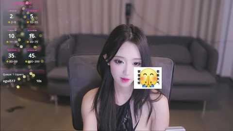 Media: Video of an East Asian woman with long black hair, wearing a black top, seated indoors. Background shows a grey sofa, Christmas tree, and digital clock overlay.