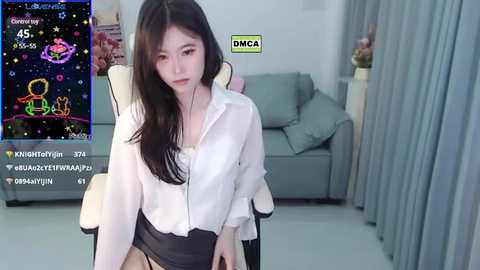 Media: Video of an East Asian woman with long black hair, wearing a white blouse and black shorts, sitting on a chair in a modern living room.